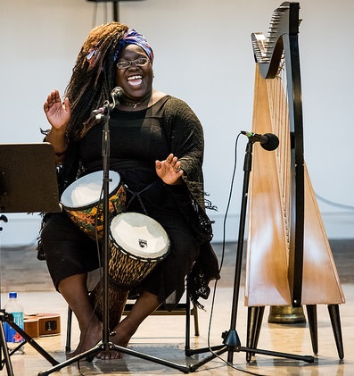Edem Garro sings behind a microphone holding two djembe drums with a harp nearby