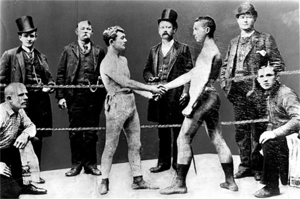 Two fighters shake hands in a boxing ring while others watch on